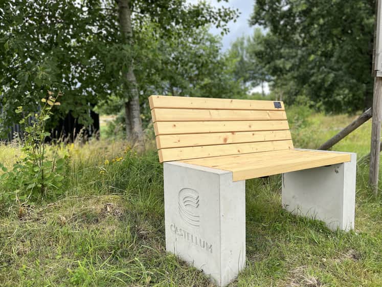 Bänk med ljusa träribbor som sits och rygg. Grå betong partier på sidorna med infräst Castellum logo.