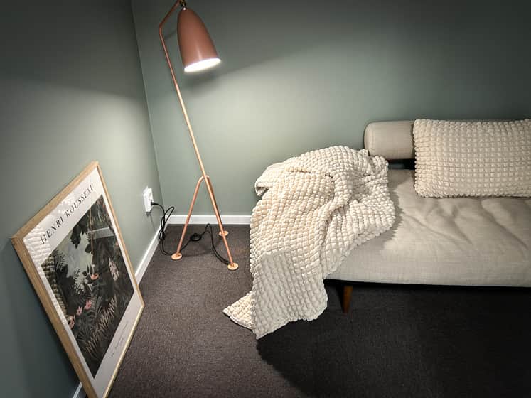 Inredning - En rosa golvlampa och en ljusgrå soffa med vit pläd och kuddar. En tavla med ljus ram står lutad mot väggen.