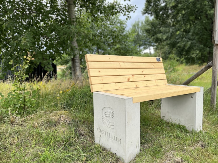 Bänk med ljusa träribbor som sits och rygg. Grå betong partier på sidorna med infräst Castellum logo.
